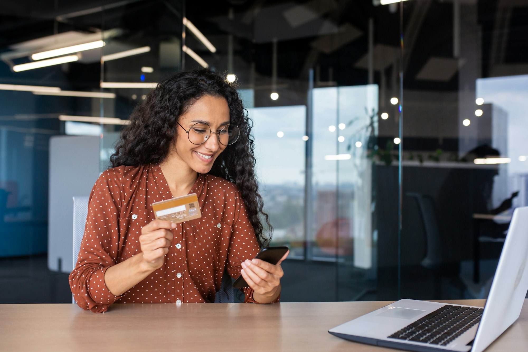 Young successful smiling woman at workplace, using app on phone and bank credit card for online